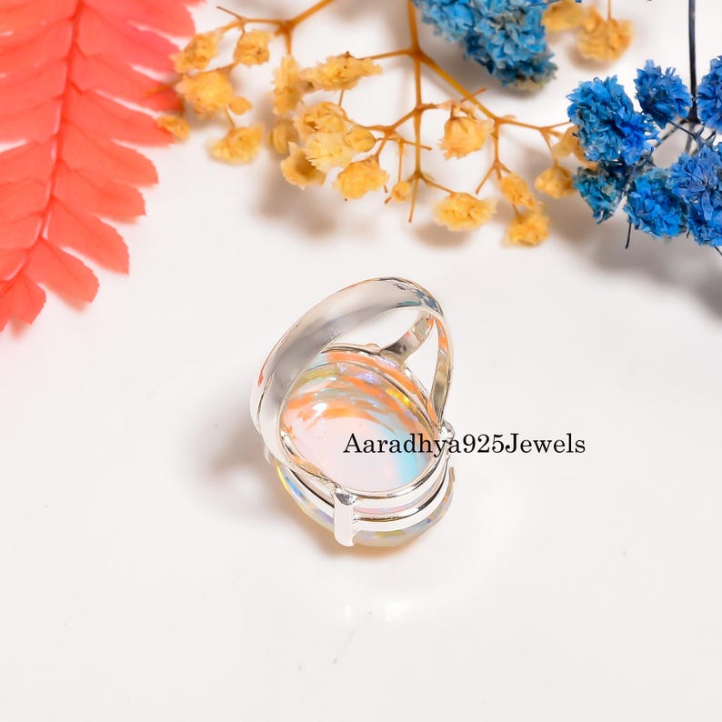May include: A silver ring with a large, oval-shaped, iridescent gemstone. The gemstone displays a mix of colors, including orange, pink, and blue. The ring is set against a white background with colorful floral accents. The text "Aaradhya925Jewels" is visible.