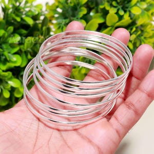 Puede incluir: Un conjunto de pulseras plateadas brillantes, que reflejan la luz, se sostiene en una mano. Las pulseras son delgadas y lisas, con un diseño simple y elegante. El fondo está borroso, con follaje verde visible.