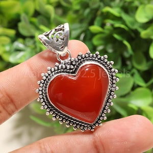 Puede incluir: Un colgante en forma de coraz&oacute;n con un centro de piedra roja oscura pulida. El colgante est&aacute; enmarcado por un borde de cuentas plateado y una fianza decorativa. El fondo es un follaje verde borroso.
