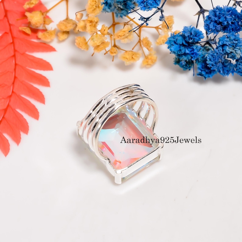 May include: A silver ring with a large, rectangular, iridescent gemstone. The ring has a multi-band design around the gemstone. The text "Aaradhya925Jewels" is visible on the ring. The background includes colorful flowers.