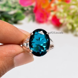 Op de afbeelding: Een zilveren ring met een grote, ovaal geslepen, diepblauwe edelsteen vastgehouden door vier pootjes. De ringband heeft een gespleten ontwerp aan de zijkanten. De tekst Aaradhya925Jewels is zichtbaar bovenaan de edelsteen. Gepolijst metaal.