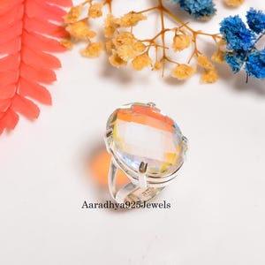 May include: A large, oval-cut gemstone ring with a rainbow iridescent effect. The ring has a silver band and setting. The text "Aaradhya925Jewels" is visible on the ring. The background includes colorful flowers.