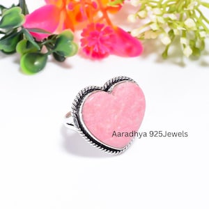 Peut inclure: Une bague en forme de cœur avec une pierre rose lisse sertie dans une bande en métal argenté torsadé. Le texte Aaradhya 925Jewels est imprimé sur le côté droit de la bague. Le design inclut une bordure argentée en corde autour du cœur rose.