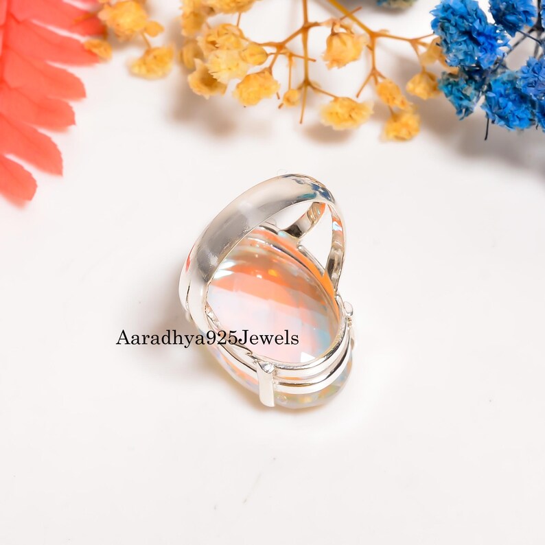 May include: A silver ring with a large, oval-shaped gemstone. The gemstone has a mix of colors, including orange, pink, and white. The ring is set against a white background with colorful flowers. Text on the image reads "Aaradhya925Jewels."