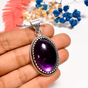 Puede incluir: Colgante ovalado de amatista con engaste plateado. El colgante tiene una piedra preciosa de color púrpura intenso y un borde texturizado. El texto "Aaradhya925Jewels" es visible. El colgante se sostiene en una mano.