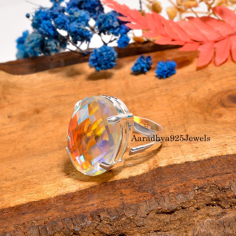 May include: A silver ring with a large, faceted, oval-shaped gemstone that reflects a spectrum of colors. The ring is set on a wooden surface with blue and red floral accents. The text "Aaradhya925Jewels" is visible on the ring.
