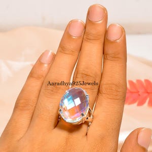 May include: A large, oval-cut gemstone ring with a silver band. The gemstone displays a rainbow of colors, reflecting light. The ring is worn on a finger, showcasing the jewelry's design. The text "Aaradhya925Jewels" is visible.