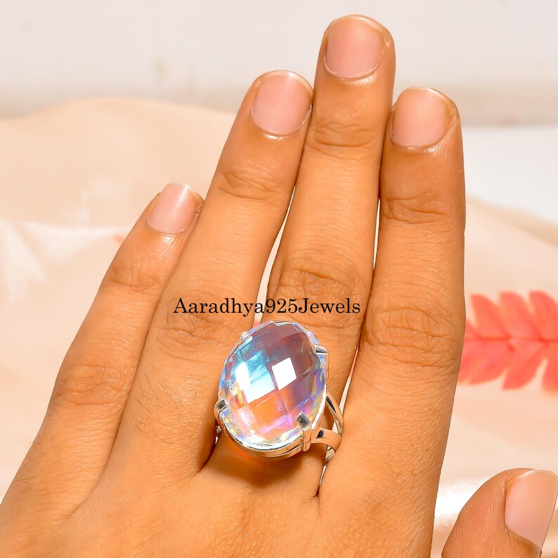 May include: A large, oval-cut gemstone ring with a silver band. The gemstone displays a rainbow of colors, reflecting light. The ring is worn on a finger, showcasing the jewelry's design. The text "Aaradhya925Jewels" is visible.