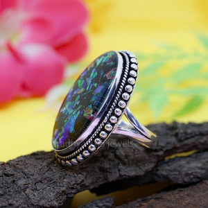 May include: A silver ring with an oval, iridescent black opal stone. The stone is framed by a beaded border and set on a silver band. The ring is displayed on a piece of dark wood against a yellow background.