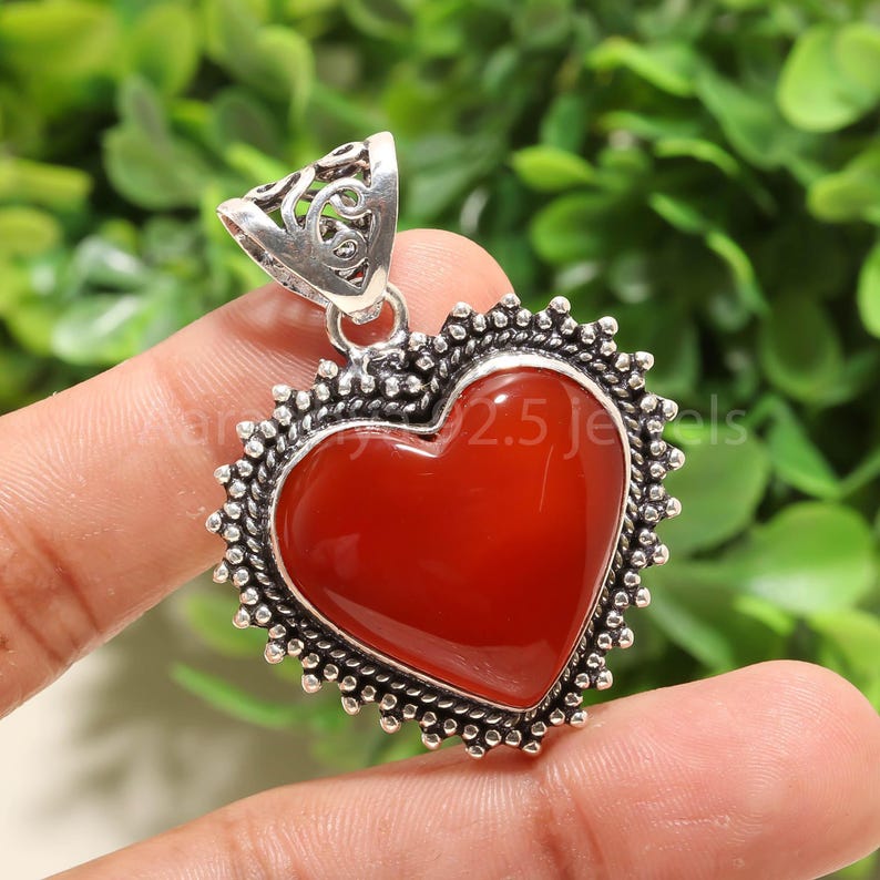 Puede incluir: Un colgante en forma de coraz&oacute;n con un centro de piedra rojo oscuro pulido. El colgante est&aacute; enmarcado por un borde de cuentas plateado y una fianza decorativa. El fondo es follaje verde borroso.
