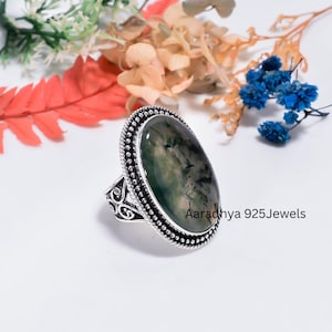 May include: An oval moss agate ring set in a silver band with a beaded border and intricate scrollwork on the sides. The stone features green inclusions resembling foliage. The text Aaradhya 925Jewels is printed in black on the lower right side.