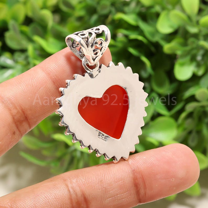 Puede incluir: Un colgante en forma de coraz&oacute;n con un centro rojo y un borde plateado texturizado. El colgante tiene una fianza decorativa y est&aacute; sostenido por los dedos de una persona. El texto "Aar ya 92.5 wels" es visible en el colgante.