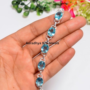 May include: A silver bracelet featuring six oval-cut, light blue gemstones linked together by small silver loops. The text Aaradhya 925Jewels is printed across the center of the bracelet. The metal links are polished and reflective.