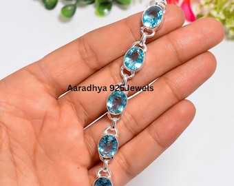 Puede incluir: Una pulsera de plata con seis piedras preciosas de color azul claro y corte ovalado, unidas por pequeños eslabones de plata. El texto Aaradhya 925Jewels está impreso en el centro de la pulsera. Los eslabones metálicos están pulidos.