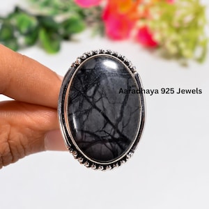 May include: An oval ring featuring a polished black and grey stone with intricate dark veining, set in a decorative silver metal band with a beaded edge. The text Aaradhaya 925 Jewels is printed on the right side of the stone surface.