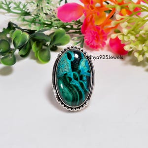 Puede incluir: Un anillo ovalado con una piedra preciosa de malaquita y crisocola pulida con patrones de remolinos en verde oscuro y turquesa vibrante. La piedra está engastada en un bisel de plata con borde de cuentas, con el texto Aaradhya925Jewels.