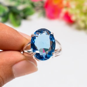 Può includere: Un anello in argento con una grande pietra preziosa blu dal taglio ovale e sfaccettato, tenuta in posizione da quattro griffe. Il gambo dell'anello è diviso in due sottili segmenti in argento lucido che si uniscono alla base della montatura.