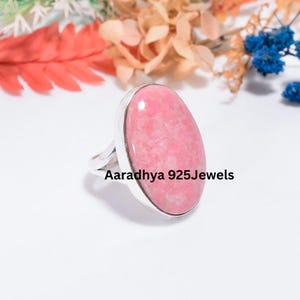 May include: A sterling silver ring featuring a large, oval-shaped, polished pink rhodochrosite gemstone set in a smooth metal bezel. The band is made of polished silver. The text Aaradhya 925Jewels is printed on the image.