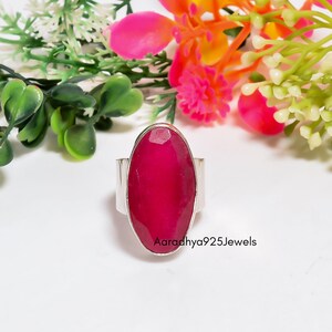 May include: A silver ring featuring a large, oval-shaped, faceted deep pink gemstone set in a smooth metal bezel. The band is plain and metallic. The text Aaradhya925Jewels is printed in black on the lower right side of the ring setting.