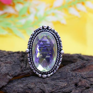 May include: Oval-shaped silver ring with a large, faceted, light purple gemstone. The ring has a detailed, braided border and small decorative accents. The gemstone reflects light, and the ring is set against a dark wood surface.