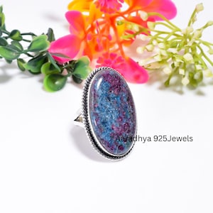 May include: An oval-shaped ring featuring a speckled blue and magenta gemstone set in a silver frame with a twisted rope border. The text Aaradhya 925Jewels is visible on the right side of the ring. The gemstone has a textured appearance.