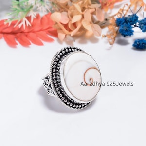 May include: A silver ring featuring a round, white Shiva eye shell with a natural brown spiral pattern. The setting is crafted with a beaded border and intricate scrollwork on the sides. The text Aaradhya 925Jewels is visible on the shell surface.
