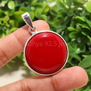 May include: A circular, red pendant with a silver-coloured bail. The pendant has a smooth, polished surface and is set in a silver-coloured bezel. The text "Aardhya 92.5 jewels" is visible on the pendant. The background is blurred green foliage.