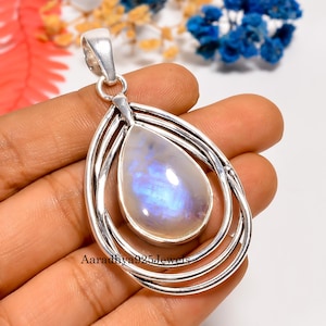 May include: A silver pendant with a teardrop-shaped moonstone. The moonstone has a pearlescent, iridescent quality with blue and white hues. The pendant is suspended within a series of concentric oval frames. The text "Aaradhya925Jewels" is visible.