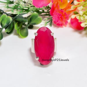 May include: A silver ring featuring a large, oval-shaped, faceted hot pink gemstone held by four prongs. The band is smooth and metallic. The text Aaradhya925Jewels is printed in black near the base of the gemstone on the white surface.