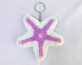 May include: A five-pointed starfish keychain crafted from white and lavender yarn. The center and each arm tip are accented with small, round, white faux pearls. A silver-toned metal split ring is attached to the top of the crocheted starfish.