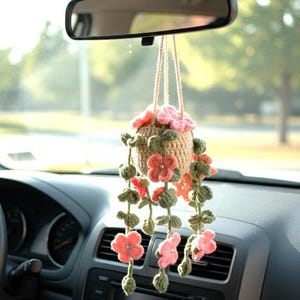 May include: A handmade crochet car charm featuring a hanging basket filled with pink flowers and green leaves. The basket is suspended from the rearview mirror, adding a touch of floral decor to the car's interior.