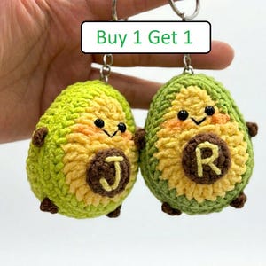 May include: Two crocheted avocado keychains with green outer skins and yellow centers. Each avocado features a brown pit embroidered with the letters J and R respectively, black bead eyes, and a small sign above reading Buy 1 Get 1.