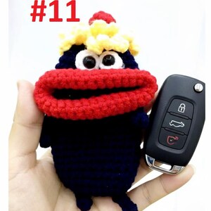 May include: A handmade, blue, red, and yellow crocheted key cover shaped like a cartoon character, with large white eyes and a red mouth. The cover is held next to a black car key. The number "#11" is in red in the upper left corner.