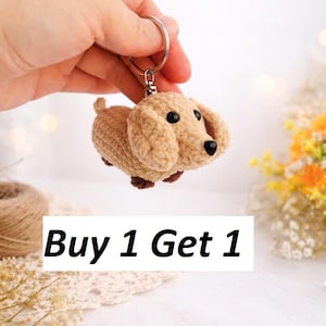 Puede incluir: Un llavero de perro salchicha tejido a ganchillo en color beige con ojos y nariz negros. El dije en forma de perro tiene un llavero plateado. La imagen incluye el texto "Buy 1 Get 1".
