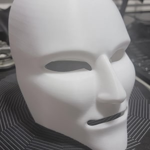 Könnte beinhalten: Eine weiße, 3D-gedruckte Maske mit glatter Oberfläche. Die Maske hat Augen- und Mundöffnungen sowie eine markante Nase. Sie liegt auf einer schwarz-silbernen, gemusterten Oberfläche, was auf einen Dekorations- oder Kostümartikel hindeutet.