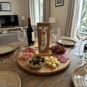 Peut inclure: Une table en bois dressée pour un apéritif, avec une planche de service ronde en bois garnie de salami, de fromage, d'olives et de raisins. Une bouteille de vin rouge, des verres à vin et une pancarte en bois avec "Rdv Apéro" sont également présents.