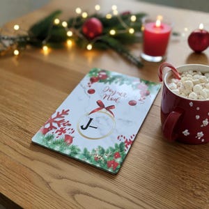 Peut inclure: Une scène de Noël festive avec une tasse rouge remplie de guimauves et une canne en sucre. Une carte de vœux avec le texte "Joyeux Noël" est posée sur une surface en bois. Des bougies et des ornements rouges complètent le décor de fête.