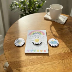 Peut inclure: Sur une table en bois, un ensemble de boutons sur le thème de l'humeur et une carte avec du texte en français. Les boutons présentent des illustrations d'un soleil, de nuages et de pluie. La carte a un motif arc-en-ciel et le texte "La météo de mon humeur du jour".