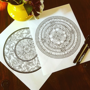 May include: Two detailed black and white mandala designs on white paper. One is a full circle, the other a half-circle. A yellow vase with flowers is in the background, with two black pens.