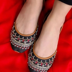 May include: A pair of black flats with intricate embroidery. The shoes feature a brown trim and are adorned with red, gold, and white embellishments. The flats are set against a vibrant red background.