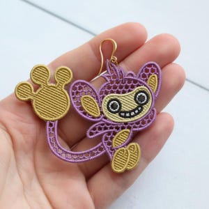 May include: A whimsical, handmade earring featuring a purple monkey design with gold accents. The monkey has a smiling face, and the earring is suspended from a gold-colored hook. The design is intricate and detailed.