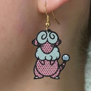 Puede incluir: Pendientes bordados de personaje de dibujos animados rosa y azul con ganchos dorados. Los pendientes presentan un diseño detallado con contornos negros y una pequeña esfera azul. Los pendientes están sujetos a un gancho dorado.