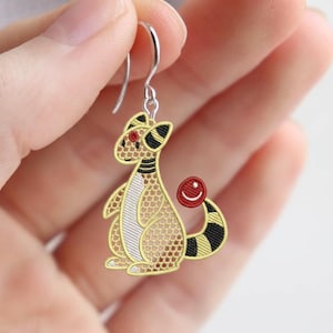 Puede incluir: Pendiente de Ampharos bordado con un gancho plateado. El pendiente presenta un diseño de Ampharos amarillo, negro y rojo. El Ampharos tiene un ojo rojo y una bola roja en la cola. El pendiente está sostenido por una mano.