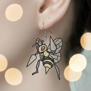 Puede incluir: Pendientes con forma de abeja. Presentan un cuerpo a rayas amarillas y negras, alas grises y un aguijón. La cabeza de la abeja es amarilla con ojos rojos. Los pendientes cuelgan de ganchos plateados.