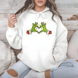 May include: White hoodie featuring a heart made of green hands with red cuffs and yellow string lights. The hands are in the shape of a heart. The hoodie has a drawstring hood and a front pocket.