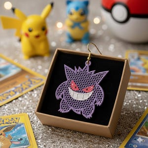 May include: A purple Gengar embroidered earring with a gold hook is displayed in a small brown box with a black interior. The earring features red eyes and a white grin. Other Pokemon-themed items are in the background.