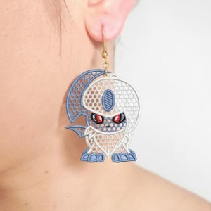 Op de afbeelding: Een goudkleurige oorbel met een geborduurd ontwerp van een stripfiguur. Het figuur heeft een wit en blauw kleurenschema met rode ogen. De oorbel hangt aan een goudkleurige haak. Het ontwerp is gedetailleerd.
