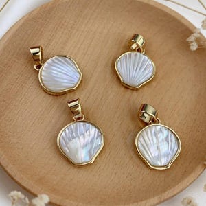 May include: Four gold-tone shell pendants with iridescent white shell inlays. The pendants are displayed on a wooden plate, with delicate white flowers in the background. Each pendant has a gold loop for a chain.