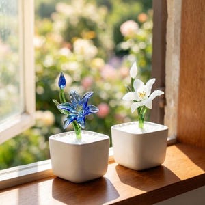 Puede incluir: Dos esculturas de flores de cristal en macetas blancas cuadradas sobre un alféizar de madera. Una flor es azul, la otra es blanca. El fondo es una vista borrosa de un jardín con follaje verde y flores rosas.