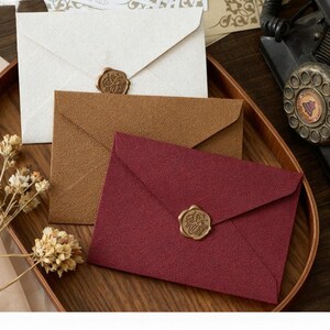 Peut inclure: Trois enveloppes texturées, crème, marron et bordeaux, chacune scellée avec un cachet de cire doré. Les enveloppes sont disposées sur un plateau en bois, suggérant un thème de papeterie ou d'invitation.